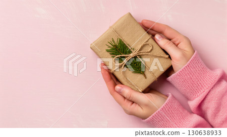 Christmas gift wrapped in kraft paper with pine twig and twine, held by hands in pink sweater on pastel pink background 130638933