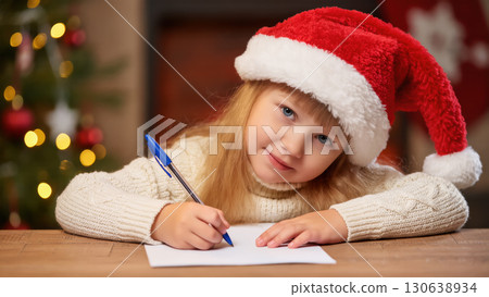 Christmas girl in Santa hat writing letter with pen on paper at wooden table, festive lights and tree in cozy background 130638934