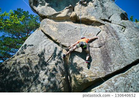 Aerial view of male rock climber ascending rugged limestone cliff with harness and rope for safety. Sportsman climbing on vertical large boulder at Dobvush Rocks in Carpathian mountains, Ukraine. 130640652