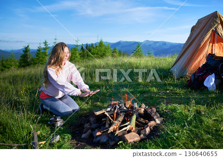 Young woman warming her hands by campfire in picturesque mountain meadow. Orange tent and camping gear. Tourist surrounded by lush greenery and distant mountains under bright blue sky. 130640655