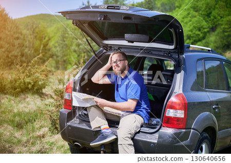 Man sits on open trunk of SUV in scenic, green landscape, holding map and scratching his head in confusion. Wearing blue shirt, beige pants, and colorful socks, he trying to figure out his location. 130640656