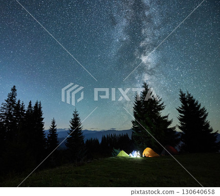 Night starry sky over grassy hill with illuminated tourist tents and coniferous trees. Mountain valley with camp tents under sky with stars and Milky Way. Hiking, night camping and astronomy concept. 130640658
