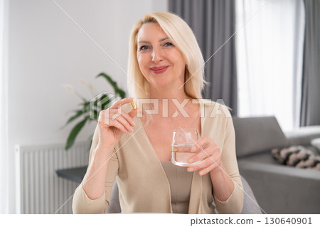 Blonde middle aged woman sitting by bright kitchen window, holding pill and glass of water, morning self care ritual, casual outfit Blonde middle aged woman sitting by bright kitchen window, holding pill and glass of water, morning self care ritual, casual outfit 130640901