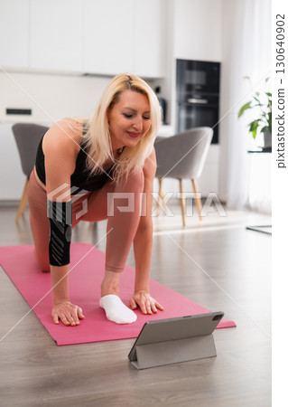Blonde woman in sportswear performing yoga pose on one knee on pink yoga mat at home, focusing on balance, posture, and stretching 130640902