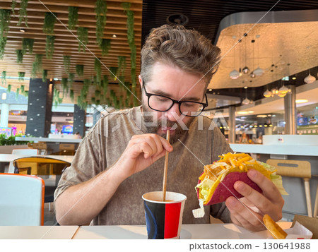 A man drinks from a cup with a straw while holding a taco at a cafe table. Consumer lifestyle, fast food culture, gastronomy and everyday dining in modern urban environment. A man drinks from a cup with a straw while holding a taco at a cafe table. Consumer lifestyle, fast food culture, gastronomy and everyday dining in modern urban environment. 130641988