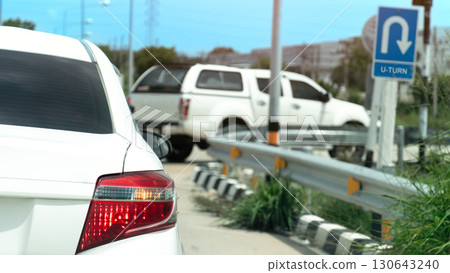 Rear side of white car with right turn signal. Open for u-turn on the road. Front of car the other car waiting for u-turn and sign board for u-turn. Background of city under blue sky. 130643240