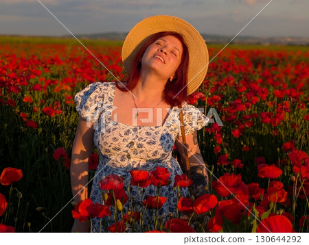 A woman smiles in a straw hat surrounded by red poppies at sunset. Lifestyle, beauty, and natural harmony of rural landscape with human presence and joy. A woman smiles in a straw hat surrounded by red poppies at sunset. Lifestyle, beauty, and natural harmony of rural landscape with human presence and joy. 130644292