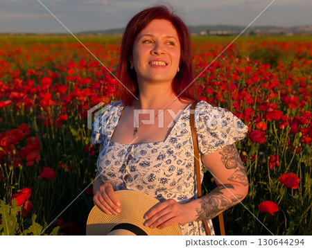 A woman smiles in a straw hat surrounded by red poppies at sunset. Lifestyle, beauty, and natural harmony of rural landscape with human presence and joy. 130644294