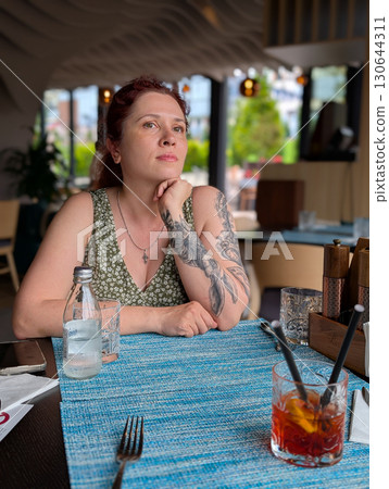 A woman sitting at a restaurant table with a cocktail. Lifestyle, dining, creativity, and relaxed urban atmosphere. A woman sitting at a restaurant table with a cocktail. Lifestyle, dining, creativity, and relaxed urban atmosphere. 130644311