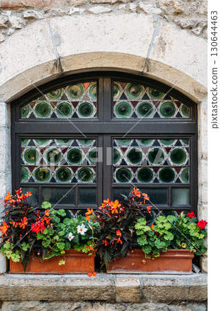 Arched window with patterned glass panes decorated by vibrant red and orange flowers in terracotta boxes. Quiet living, countryside serenity, daydream dwellings, contemplative design 130644463