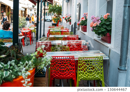 Colorful cafe terrace with red and green lattice chairs lining a narrow European street adorned with geranium flower boxes. Urban vibrancy, cafe culture, street life, pedestrian-friendly design 130644467