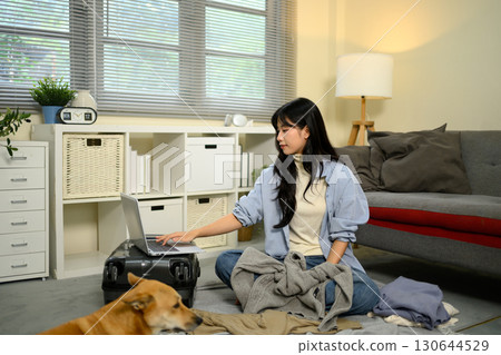 Young woman sitting on the floor using a laptop while packing clothes for travel Young woman sitting on the floor using a laptop while packing clothes for travel 130644529