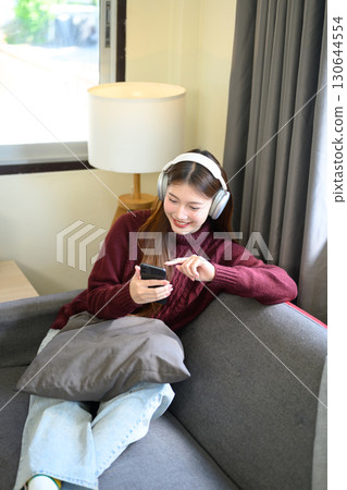 Cheerful girl in burgundy sweater listening to music and browsing phone indoors 130644554