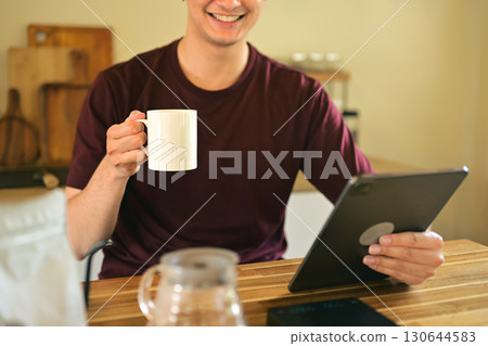 Smiling man holding a white coffee mug while using a digital tablet at home kitchen Smiling man holding a white coffee mug while using a digital tablet at home kitchen 130644583