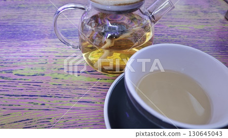 Glass teapot and a white cup filled with herbal tea on a colorful wooden table Glass teapot and a white cup filled with herbal tea on a colorful wooden table 130645043