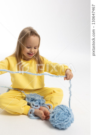 Pretty little girl in yellow clothes plays with ball of blue thread. Child model with long blond hair sits in studio on light background. High quality photo 130647467