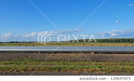 A car is driving along a highway, passing a gas pipeline that stretches across a field under a blue sky. Side view from window of a car showing a wide gas pipe along highway. A car is driving along a highway, passing a gas pipeline that stretches across a field under a blue sky. Side view from window of a car showing a wide gas pipe along highway. 130647875