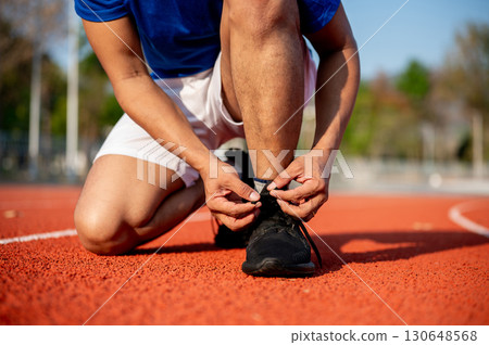 Close up of a man crouching while tying his black sneaker shoelace on racetrack. Outdoor exercise. 130648568