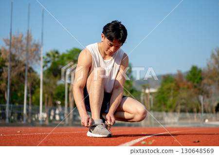An asian man in white tank top is crouching while tying his sneaker shoelace on the racetrack. 130648569