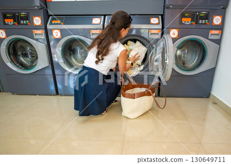 Young woman loading laundry into washing machine in self-service laundry facility. Everyday routine of washing clothes in modern public laundromat. Young woman loading laundry into washing machine in self-service laundry facility. Everyday routine of washing clothes in modern public laundromat. 130649711
