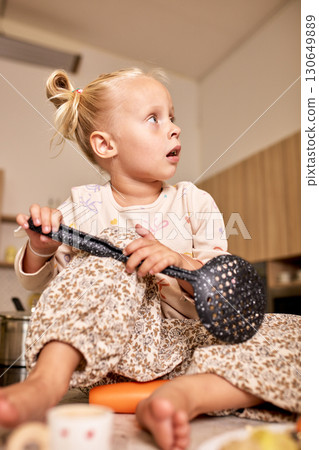 Young girl holding kitchen utensil seated on floor in a cozy kitchen 130649889