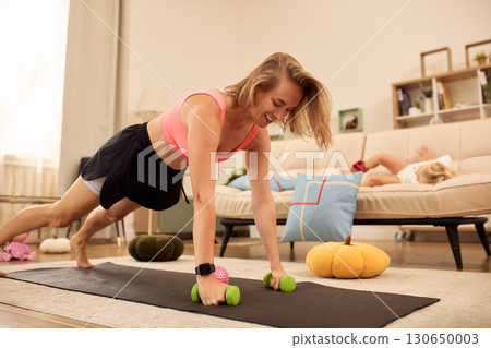 Woman exercising at home lifting dumbbells while her child rests nearby Woman exercising at home lifting dumbbells while her child rests nearby 130650003