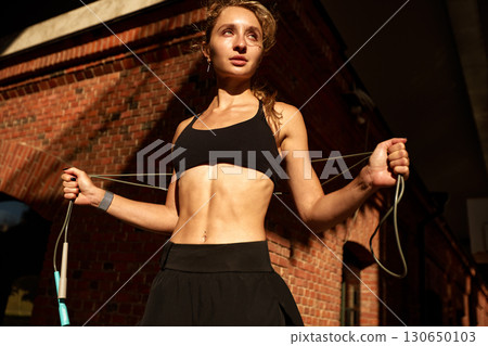 Young athletic woman jumps rope outdoors against a brick building background 130650103