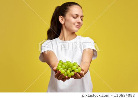 portrait of attractive Caucasian smiling woman holds grapes isolated on yellow studio background, focus on grapes portrait of attractive Caucasian smiling woman holds grapes isolated on yellow studio background, focus on grapes 130654098