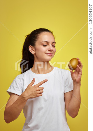 Happy woman in white t-shirt holding kiwi on studio yellow background Happy woman in white t-shirt holding kiwi on studio yellow background 130654107