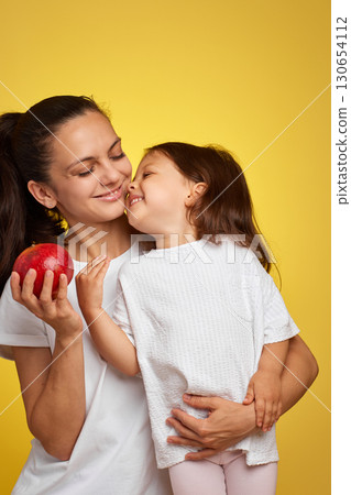 Cheerful mother and her little child girl holding pomegranate on yellow background. happy family having fun together Cheerful mother and her little child girl holding pomegranate on yellow background. happy family having fun together 130654112