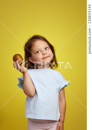 cute Caucasian little child girl holding kiwi fruit on yellow background 130654145