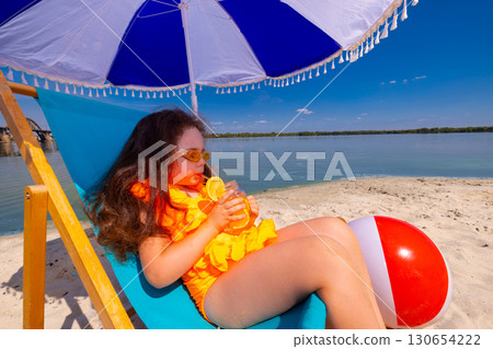 Girl in orange swimsuit sipping citrus drink under beach umbrella with beach ball 130654222