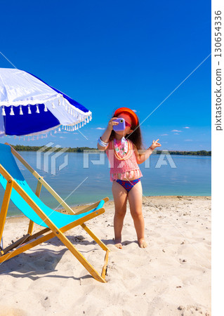 Child standing on beach near umbrella taking photo with toy camera 130654336