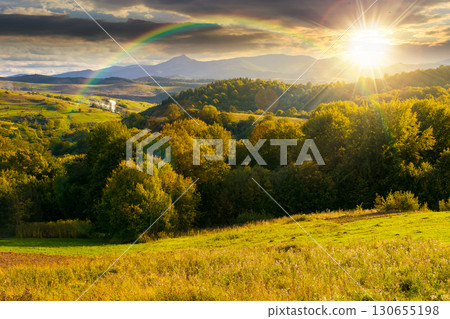 countryside landscape on september sunset. village in the valley and rural fields on the forested hills in evening light. mountain ridge with high peak in the distance under rainbow 130655198