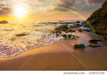 sea coast with rainbow over the boulders in the bay at sunset. sea tide with calm waves wash the golden sandy beach in evening light. beautiful autumn vacation at the seaside for honeymoon backdrop 130655199