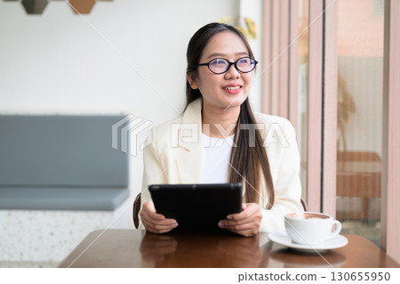 Asian cute woman sitting in coffee shop using tablet computer looking through window and thinking 130655950