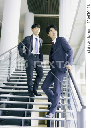 Portrait of two young businessmen standing on the stairs of an office building, vertical position Portrait of two young businessmen standing on the stairs of an office building, vertical position 130656468
