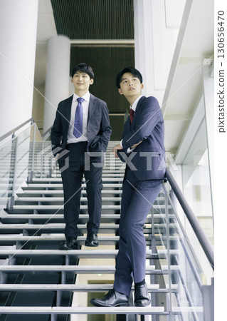 Portrait of two young businessmen standing on the stairs of an office building, vertical position Portrait of two young businessmen standing on the stairs of an office building, vertical position 130656470