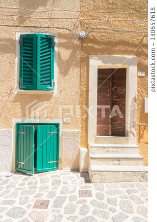 Mediterranean building facade featuring vibrant green shutters and rustic doors. The sun-drenched yellow walls and stone pavement enhance the lively, coastal ambiance of the scene. Mediterranean building facade featuring vibrant green shutters and rustic doors. The sun-drenched yellow walls and stone pavement enhance the lively, coastal ambiance of the scene. 130657618