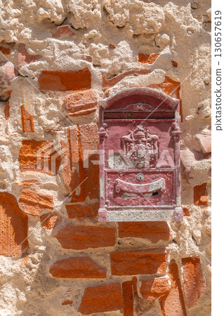 Red vintage mailbox attached to a textured brick wall, showcasing intricate details and historical charm. The weathered brick background highlights the mailboxs ornate design. Red vintage mailbox attached to a textured brick wall, showcasing intricate details and historical charm. The weathered brick background highlights the mailboxs ornate design. 130657619