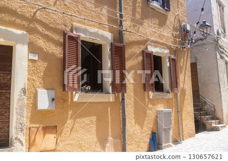 Rustic building with open windows and wooden shutters in a sunlit alley. The charming architecture features vibrant walls and classic street elements, creating a quaint, picturesque scene. Rustic building with open windows and wooden shutters in a sunlit alley. The charming architecture features vibrant walls and classic street elements, creating a quaint, picturesque scene. 130657621
