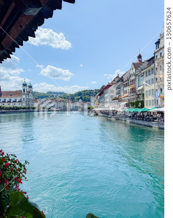 Breathtaking view of historic buildings lining a serene river under a clear blue sky. Bright flowers adorn the riverbank, while people enjoy outdoor dining by the water 130657624