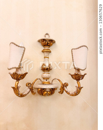 Vintage wall sconce with two lamps showcasing an elegant, ornate design. The intricate detailing in gold and white enhances its timeless appeal, perfect for classic interiors. 130657629