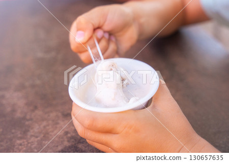Close-up of a child holding a cup of ice cream, using a small spoon to scoop it. The image captures the moment of enjoyment and focus as the child indulges in the treat. 130657635