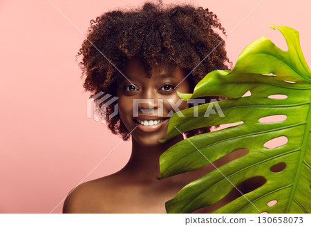African American young woman peeks out from tropical plant leaf. 130658073