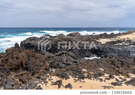 Scenic coastal view on La Graciosa island, Lanzarote, featuring volcanic rocks, sandy beach, and ocean waves under a cloudy sky. 130658668