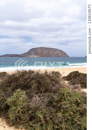 A scenic view on La Graciosa, Lanzarote, showcasing a sandy beach, the turquoise ocean, and a distant island under a cloudy sky. 130658675