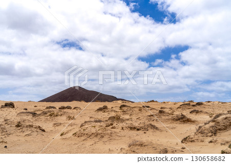 A scenic view of La Graciosa, showing a volcanic hill over sand dunes under a cloudy blue sky. 130658682