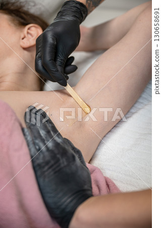 Beautician applying wax with wooden spatula on female armpit during professional hair removal procedure 130658691