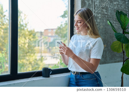 Smiling young woman using smartphone standing at home, window background Smiling young woman using smartphone standing at home, window background 130659310